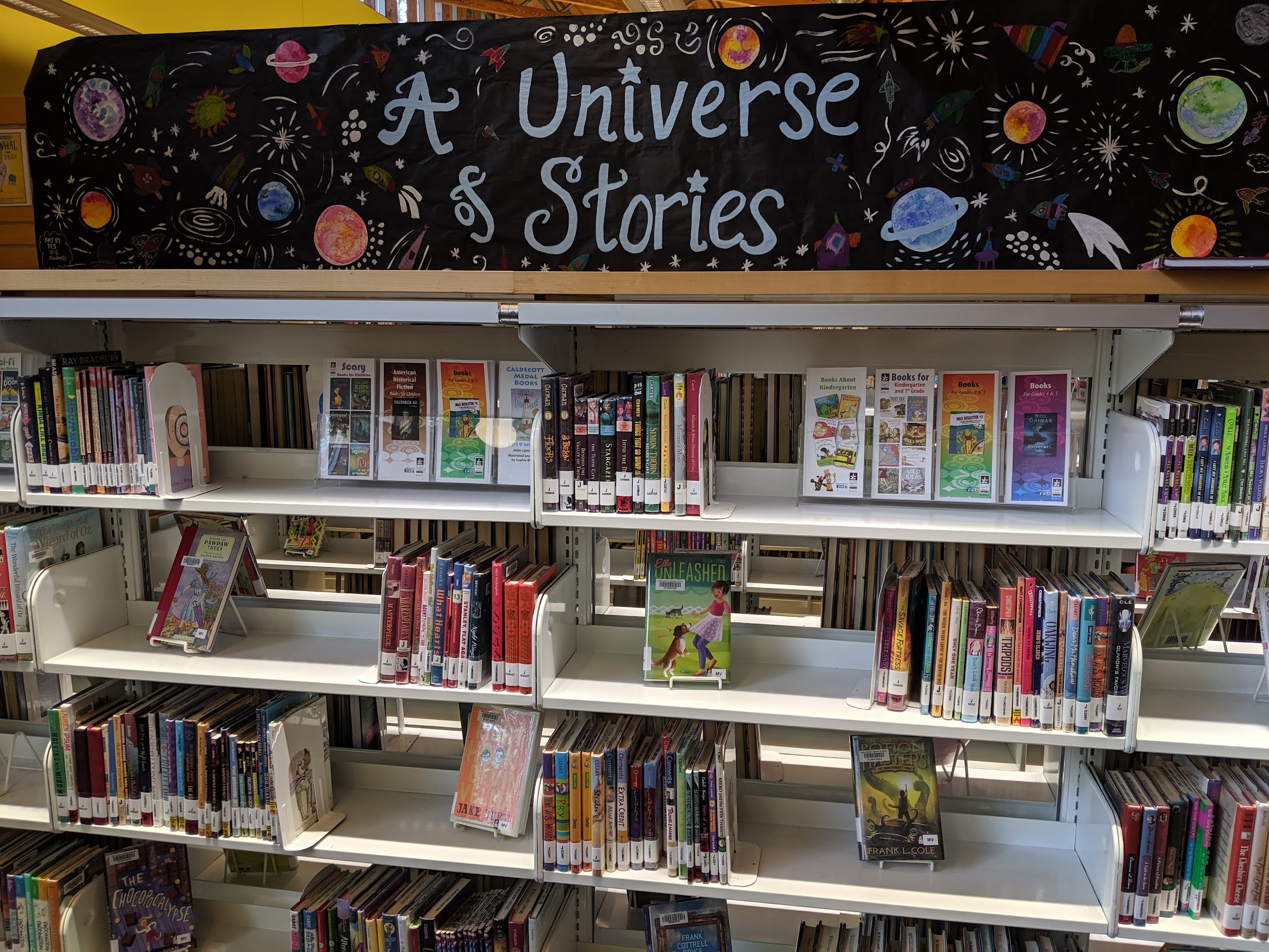 A shelf of children's books with a large sign that says "A universe of Stories" with pictures of planets and stars.